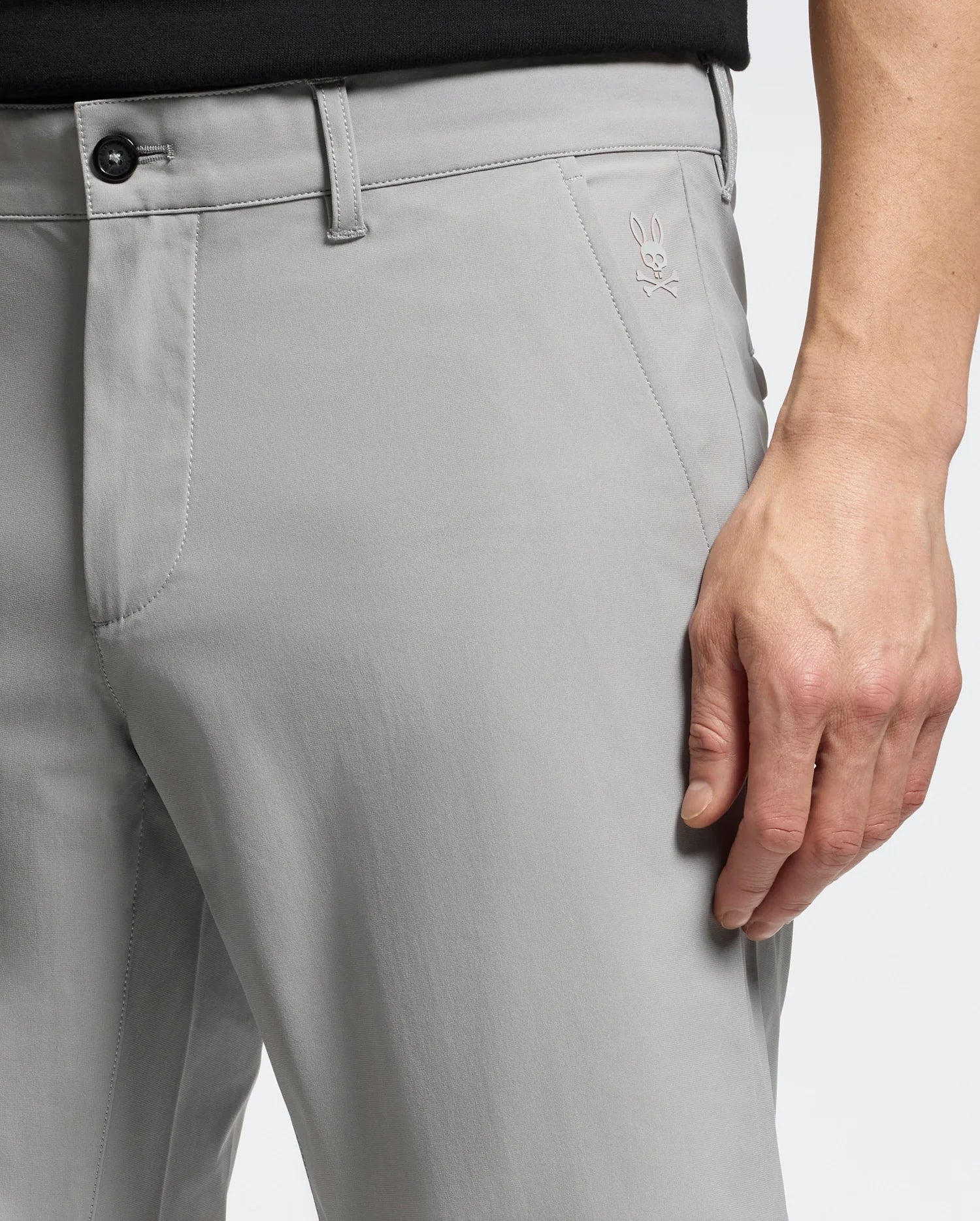 Close-up of a person in Psycho Bunny's MENS 10K STEP SHORT - B6R969F200, featuring breathable light gray fabric and a skull with rabbit ears logo on the upper thigh. They're standing with one hand relaxed at their side, wearing a black top.