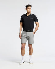 A man in a black polo shirt and Psycho Bunny's gray MENS 10K STEP SHORT - B6R969F200 stands against a plain background. He wears white sneakers and a watch, hands resting at his sides, smiling slightly, ready to enjoy the day with athletic breathability.