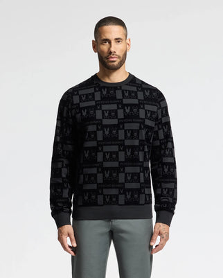 A man wearing the MENS ROMEO LIGHTWEIGHT SWEATSHIRT - B6S129E200 by Psycho Bunny, which features a stylish black crew design with the brand's signature text and logo pattern, is paired with gray pants and stands against a plain gray background.