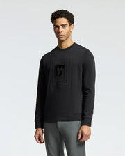 A person is wearing the Psycho Bunny MENS OAK CITY CREW NECK - B6S274E200, a premium quality black long-sleeve shirt with raised embroidery in a subtle graphic design on the chest. They are standing against a plain white background, paired with gray pants, and looking directly at the camera.
