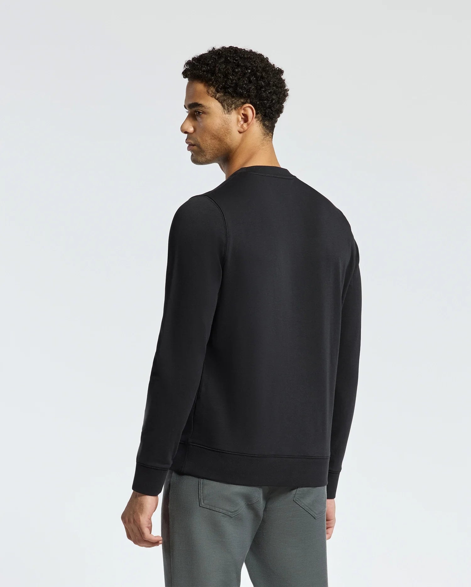 With short curly hair and their back to the camera, a person is wearing a black MENS OAK CITY CREW NECK - B6S274E200 by Psycho Bunny, featuring raised embroidery, paired with grey pants against a plain white background.