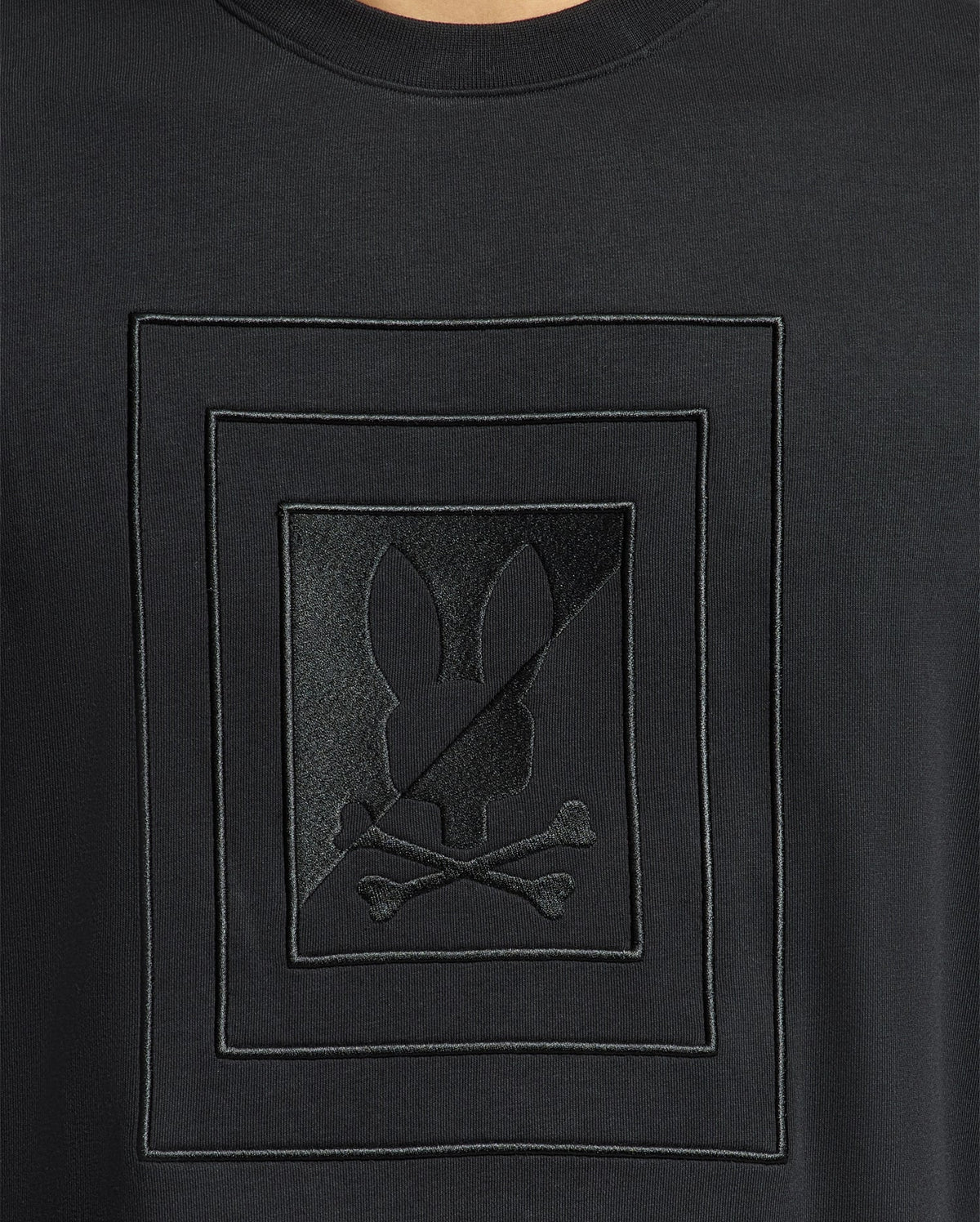 A close-up of the Psycho Bunny MENS OAK CITY CREW NECK - B6S274E200 reveals its sleek black design featuring an embossed look. At the center, a geometric pattern presents a rabbit face above crossed bones, accented by raised embroidery. This premium quality piece from Psycho Bunny showcases a glossy, textured appearance that subtly stands out against the black fabric.