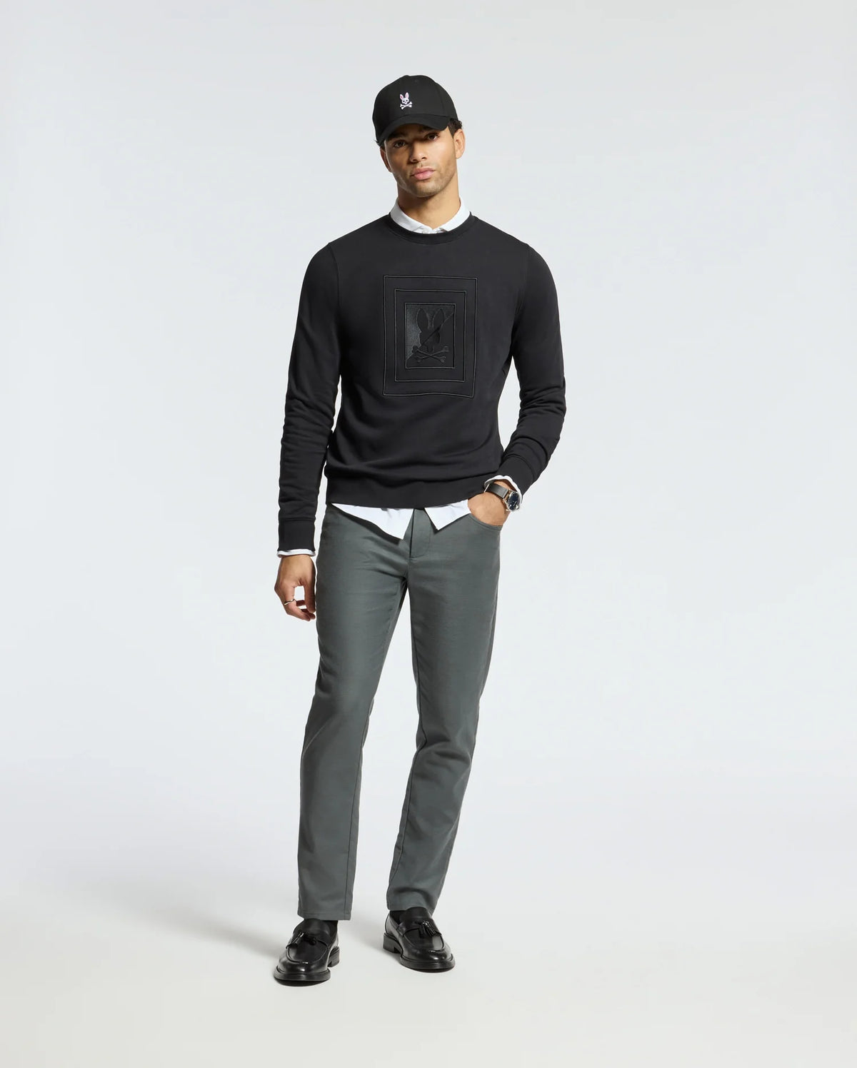 A person in a Psycho Bunny MENS OAK CITY CREW NECK - B6S274E200, styled with a white shirt and gray pants, stands against a plain background. They complete the look with black shoes and a black cap, exuding a relaxed posture.