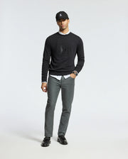 A person in a Psycho Bunny MENS OAK CITY CREW NECK - B6S274E200, styled with a white shirt and gray pants, stands against a plain background. They complete the look with black shoes and a black cap, exuding a relaxed posture.