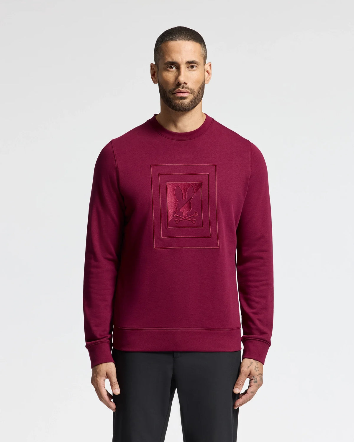 A man with short hair and a beard wears the Psycho Bunny MENS OAK CITY CREW NECK - B6S274E200 in burgundy, showcasing raised embroidery. The premium sweatshirt perfectly complements his black pants as he stands against a plain white background.