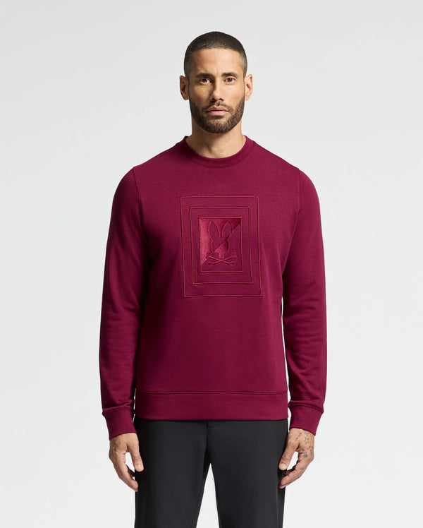 A man with short hair and a beard wears the Psycho Bunny MENS OAK CITY CREW NECK - B6S274E200 in burgundy, showcasing raised embroidery. The premium sweatshirt perfectly complements his black pants as he stands against a plain white background.