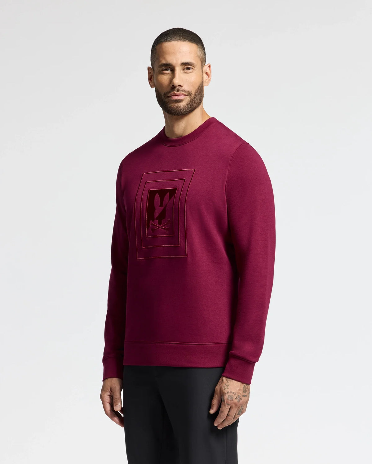 Against a white backdrop, a man models the Psycho Bunny MENS OAK CITY CREW NECK - B6S274E200 in maroon. Featuring subtle chest embroidery, his short hair and beard complement the sweater's stylish yet understated appeal.