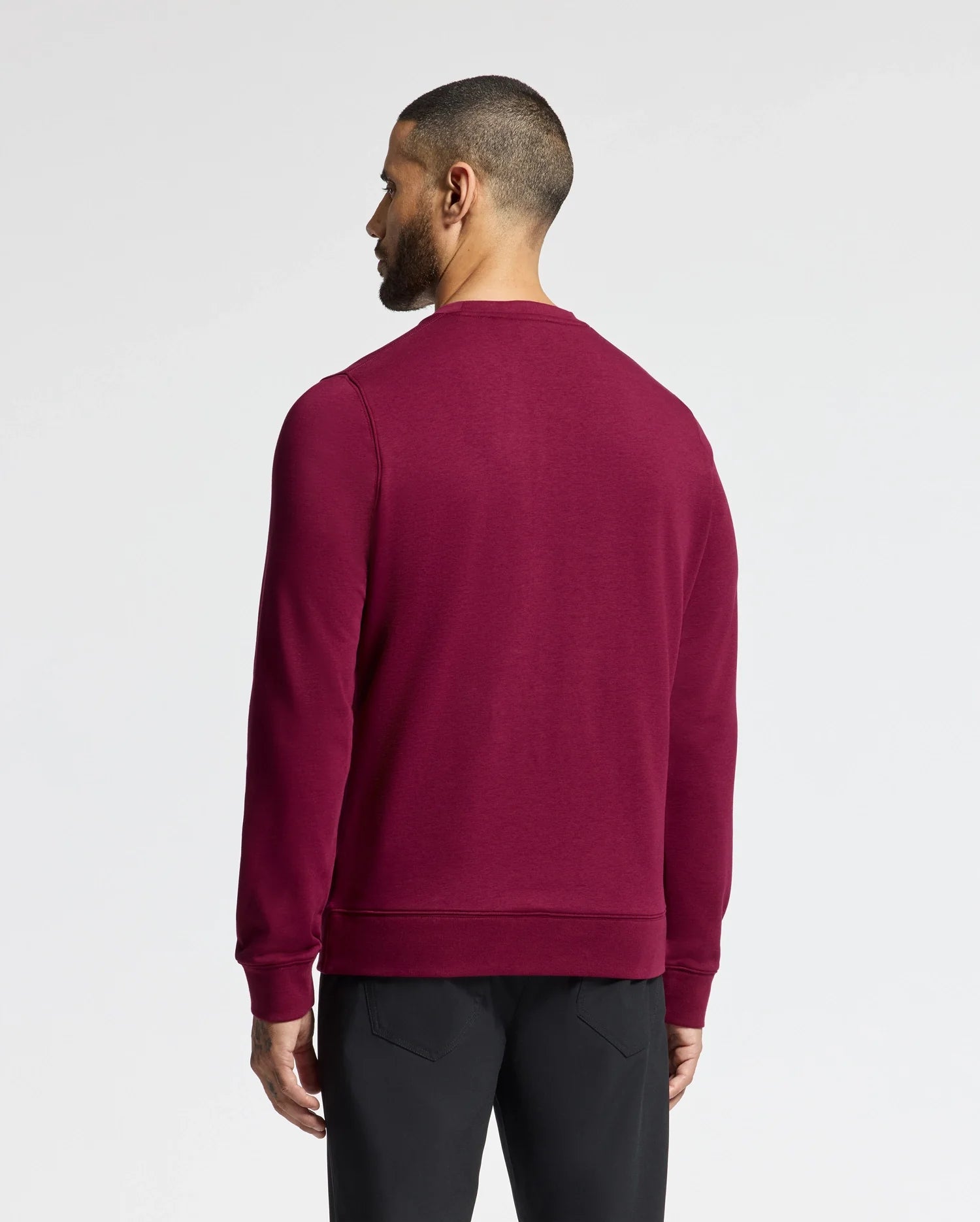 A man with short hair, facing away, wears the Psycho Bunny MENS OAK CITY CREW NECK - B6S274E200 sweater in burgundy paired with black pants. The lightweight design highlights his style against the plain white background.