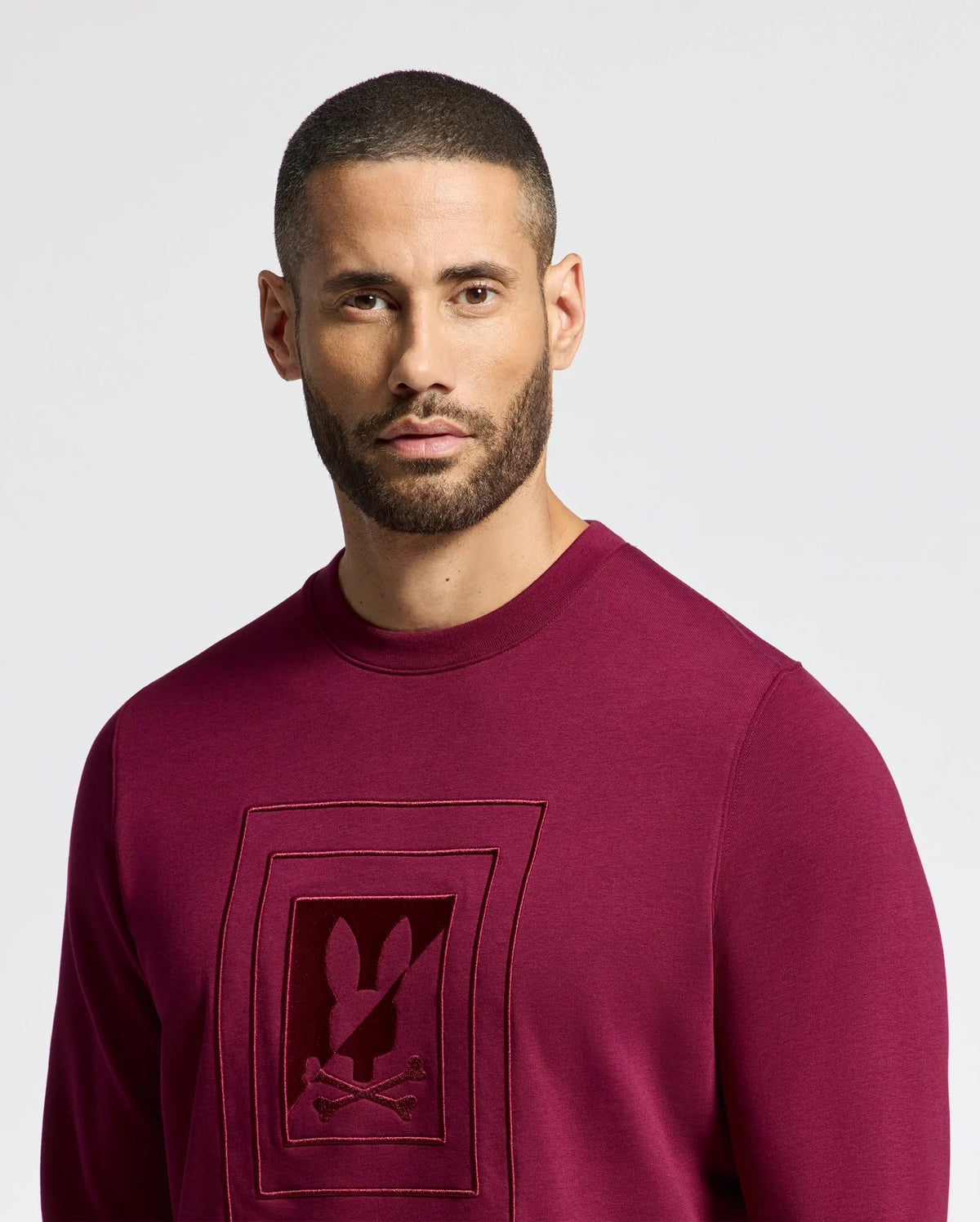 A man with a short beard and buzz cut wears a Psycho Bunny MENS OAK CITY CREW NECK - B6S274E200, featuring a rabbit logo with raised embroidery. He looks intently at the camera against a plain white background, highlighting the sweatshirt's premium quality.