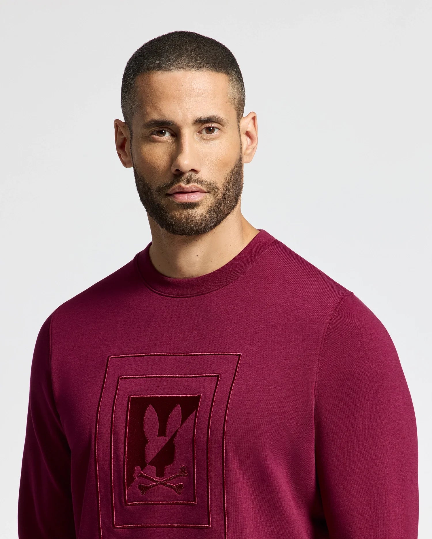 A man with a short beard and buzz cut wears a Psycho Bunny MENS OAK CITY CREW NECK - B6S274E200, featuring a rabbit logo with raised embroidery. He looks intently at the camera against a plain white background, highlighting the sweatshirt's premium quality.