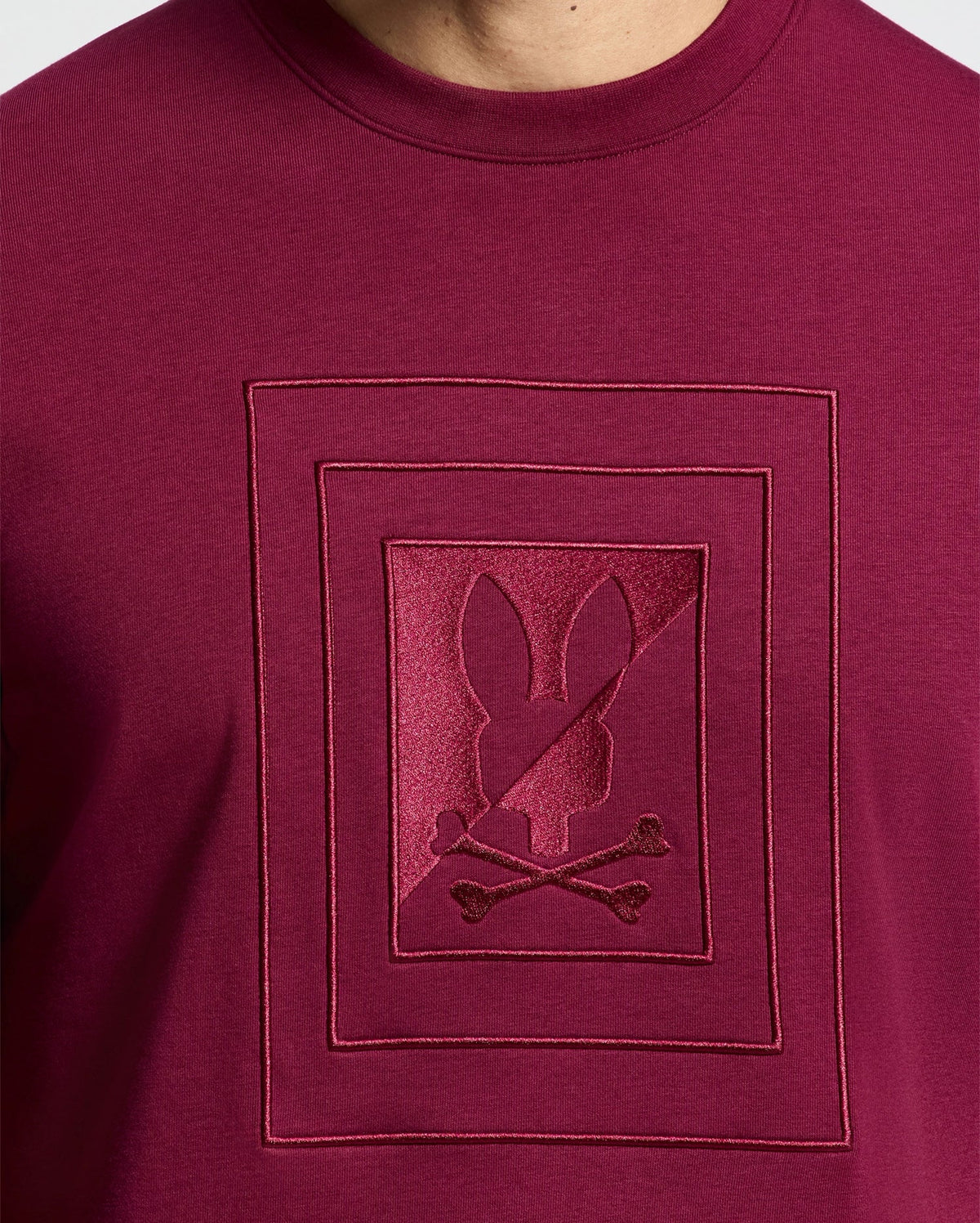 Someone models the Psycho Bunny MENS OAK CITY CREW NECK - B6S274E200, featuring a dark red hue with a central bunny skull and crossbones design, showcasing raised embroidery.