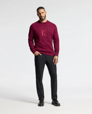 A man in a Psycho Bunny MENS OAK CITY CREW NECK - B6S274E200 sweater in burgundy with front raised embroidery stands against a white backdrop. He pairs it with black trousers and shoes. His short hair and tattoos on his left hand add a unique touch.