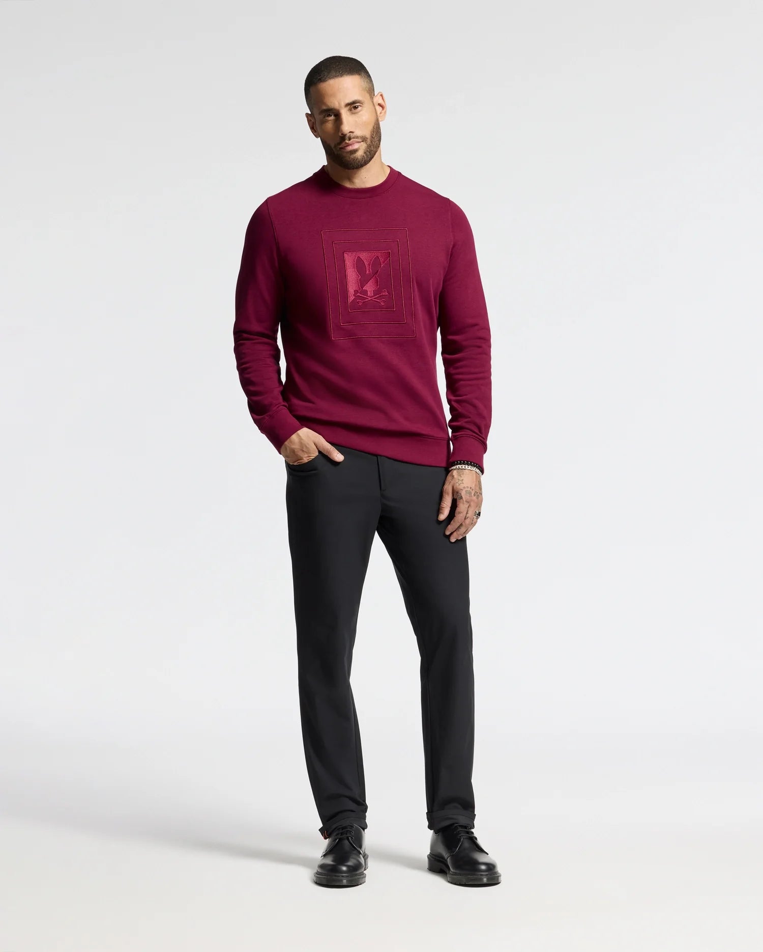 A man in a Psycho Bunny MENS OAK CITY CREW NECK - B6S274E200 sweater in burgundy with front raised embroidery stands against a white backdrop. He pairs it with black trousers and shoes. His short hair and tattoos on his left hand add a unique touch.