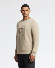 A man wearing the Psycho Bunny MENS OAK CITY CREW NECK (B6S274E200) in beige, with raised embroidery on the front, stands against a plain white background. He has short dark hair and is looking directly at the camera, paired with dark pants for a sleek look.