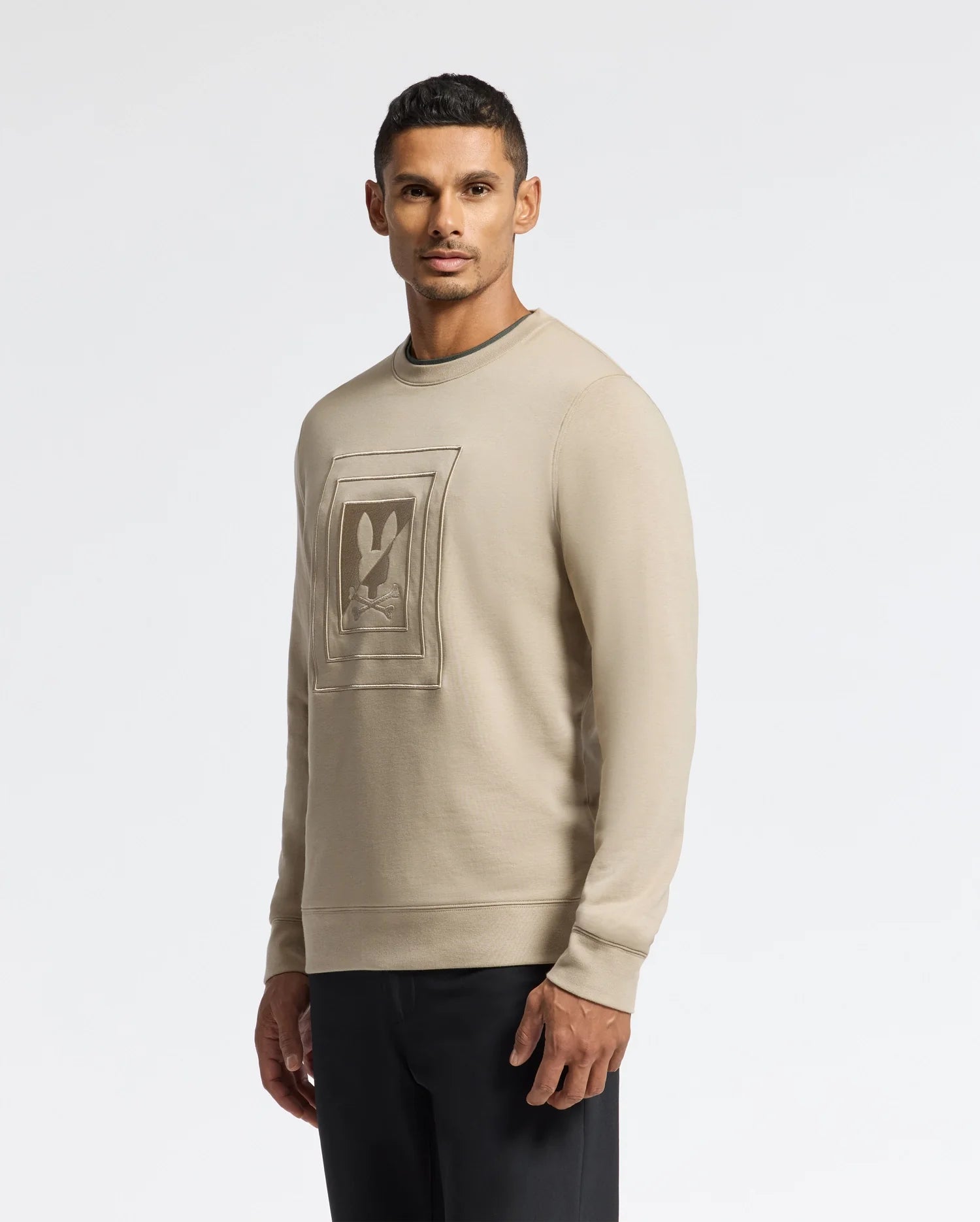 A man wearing the Psycho Bunny MENS OAK CITY CREW NECK (B6S274E200) in beige, with raised embroidery on the front, stands against a plain white background. He has short dark hair and is looking directly at the camera, paired with dark pants for a sleek look.