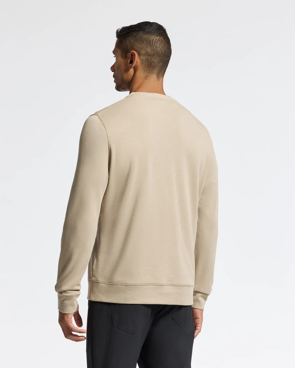 A man in a beige long-sleeve shirt and black pants stands with his back to the camera against a simple white backdrop, quietly highlighting the stylish appeal of the Psycho Bunny MENS OAK CITY CREW NECK - B6S274E200.