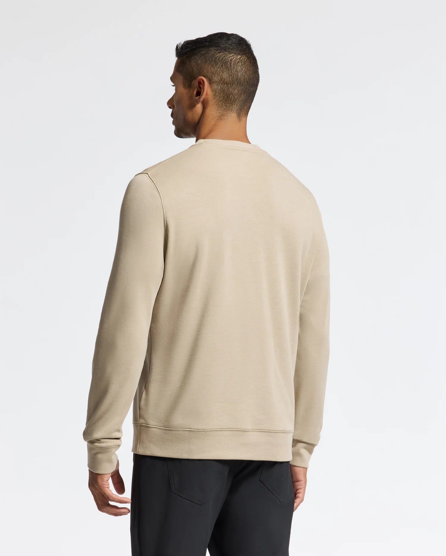 A man in a beige long-sleeve shirt and black pants stands with his back to the camera against a simple white backdrop, quietly highlighting the stylish appeal of the Psycho Bunny MENS OAK CITY CREW NECK - B6S274E200.