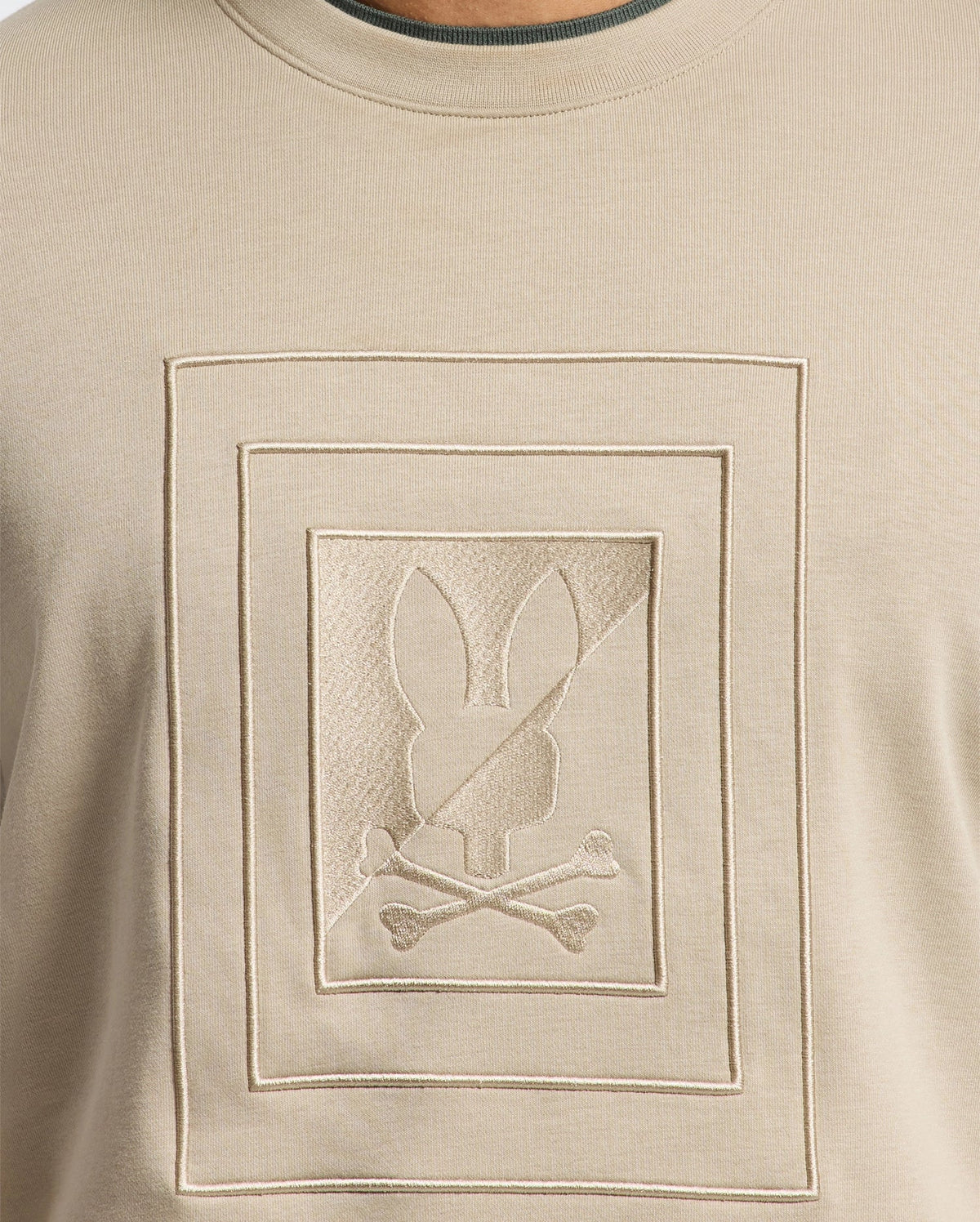 This beige MENS OAK CITY CREW NECK - B6S274E200 from Psycho Bunny features raised embroidery of a bunny silhouette above two crossed bones, all within a double-lined rectangular frame. Combining style and comfort, this lightweight men’s crew neck sweatshirt is an eye-catching addition to any wardrobe.