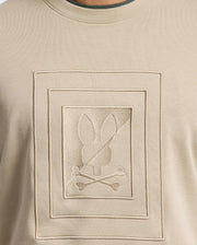 This beige MENS OAK CITY CREW NECK - B6S274E200 from Psycho Bunny features raised embroidery of a bunny silhouette above two crossed bones, all within a double-lined rectangular frame. Combining style and comfort, this lightweight men’s crew neck sweatshirt is an eye-catching addition to any wardrobe.