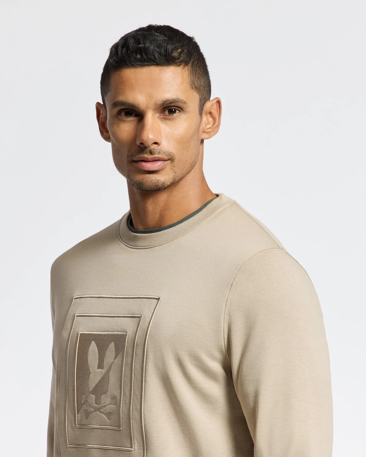 A man with short dark hair is wearing the Psycho Bunny MENS OAK CITY CREW NECK - B6S274E200 in beige, which features raised embroidery on the front. The lightweight design provides comfort and style against a plain white background.