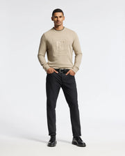 A man in a beige Psycho Bunny MENS OAK CITY CREW NECK sweater, featuring a geometric design, stands against a plain background, wearing black pants. He completes his look with black shoes and a belt, his hands casually in his pockets.