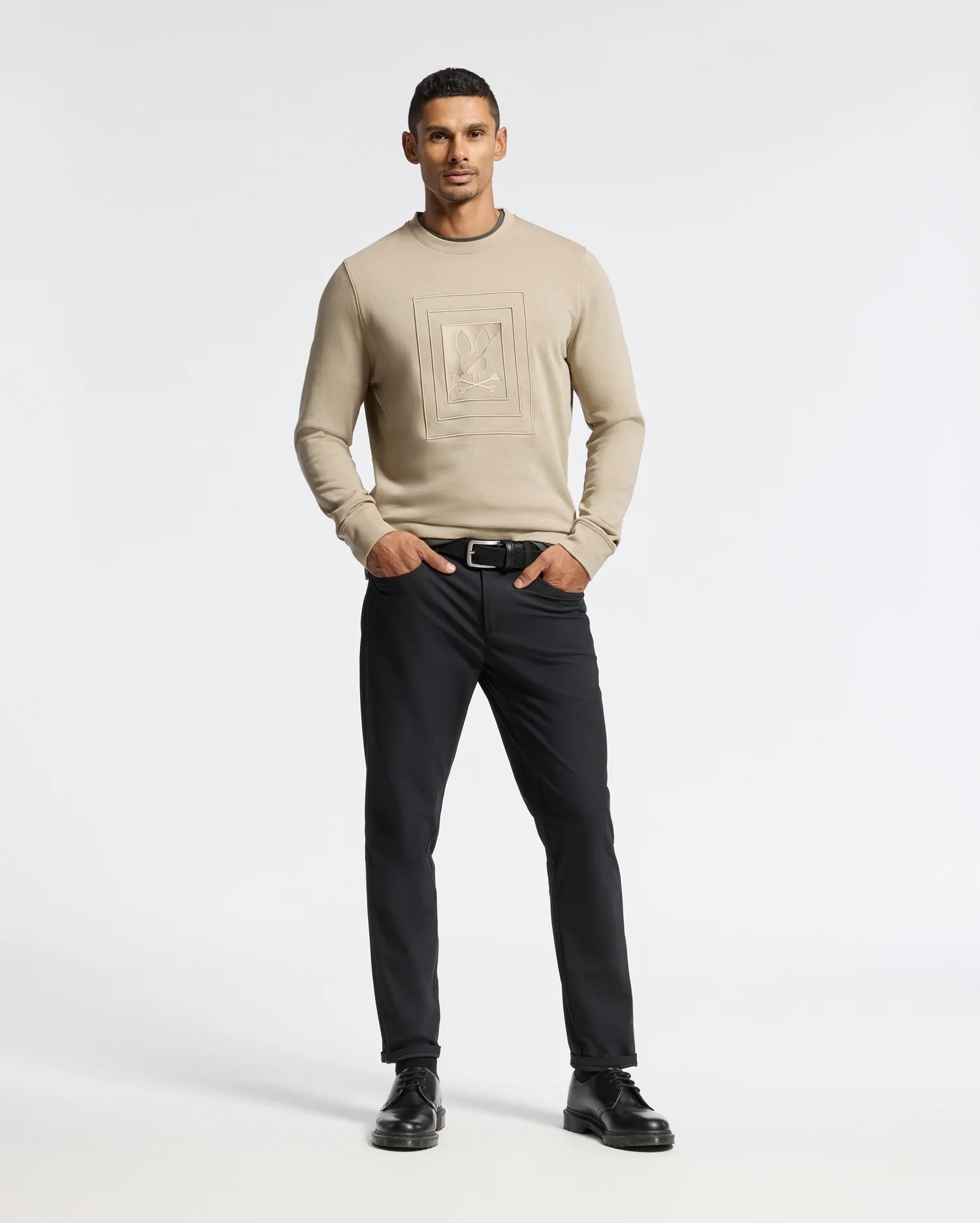 A man in a beige Psycho Bunny MENS OAK CITY CREW NECK sweater, featuring a geometric design, stands against a plain background, wearing black pants. He completes his look with black shoes and a belt, his hands casually in his pockets.