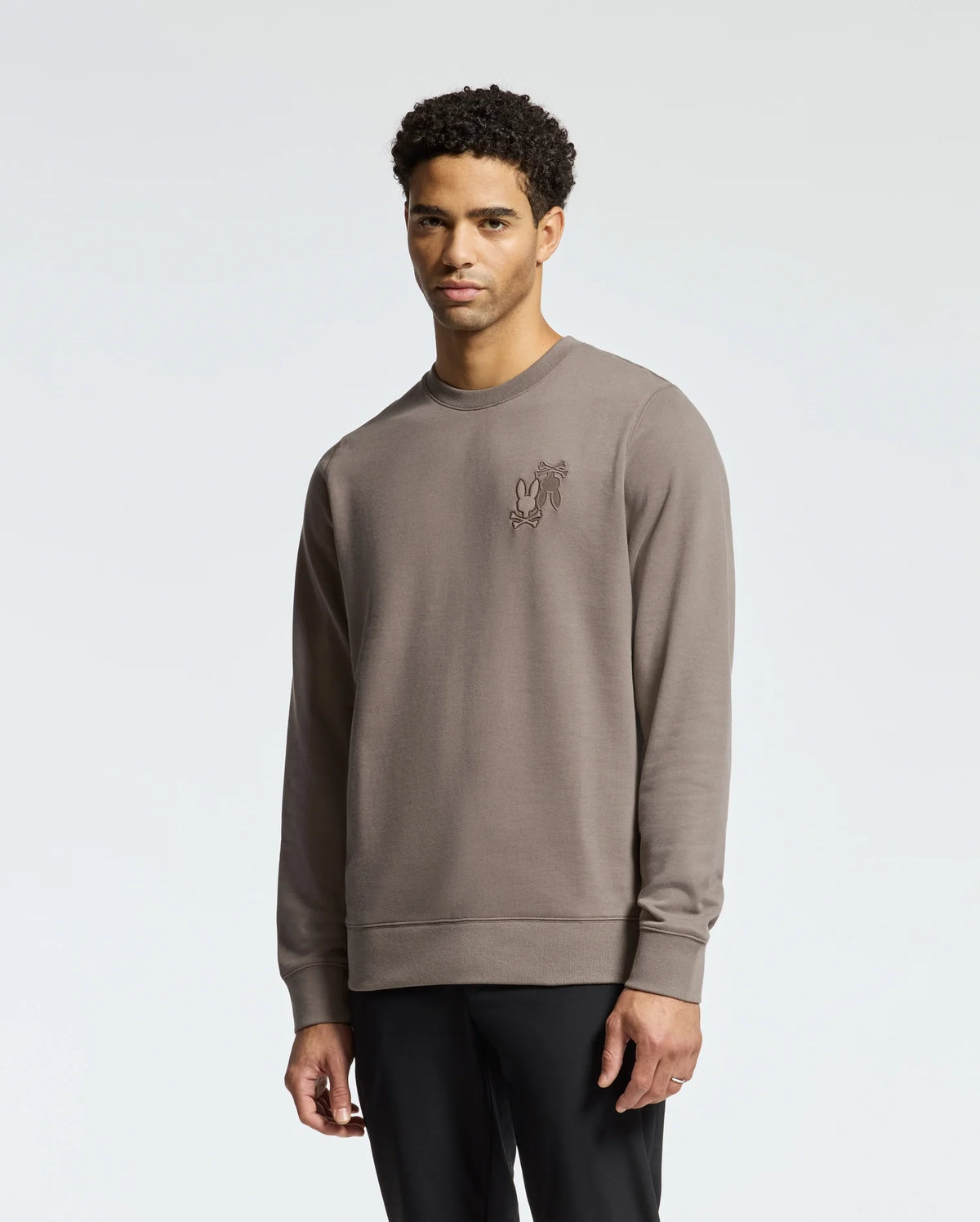 Wearing the Psycho Bunny Men's Pierce Peached Terry Crew (B6S278E200), a person with short curly hair stands against a plain background. The taupe embroidered cotton sweatshirt exudes effortless style, perfectly paired with dark pants for a modern, fashionable look.