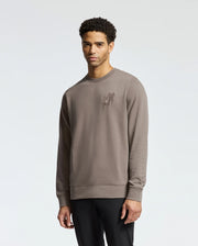Wearing the Psycho Bunny Men's Pierce Peached Terry Crew (B6S278E200), a person with short curly hair stands against a plain background. The taupe embroidered cotton sweatshirt exudes effortless style, perfectly paired with dark pants for a modern, fashionable look.