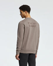 A person with short curly hair faces away, wearing the taupe MENS PIERCE PEACHED TERRY CREW - B6S278E200 by Psycho Bunny. It's made of high-quality cotton and features "Psycho Bunny" on the back, perfectly matching their dark pants against a light background.