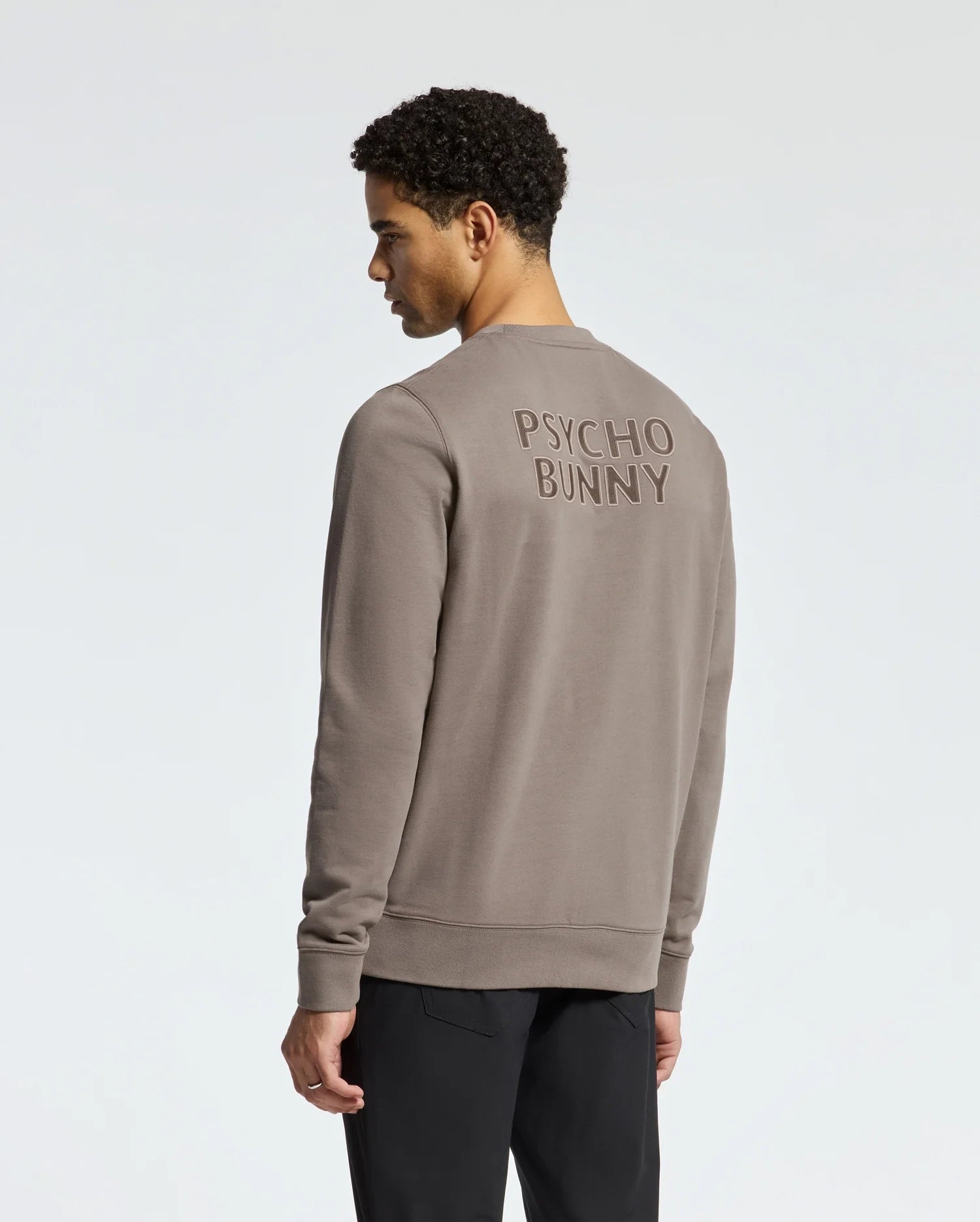 A person with short curly hair faces away, wearing the taupe MENS PIERCE PEACHED TERRY CREW - B6S278E200 by Psycho Bunny. It's made of high-quality cotton and features "Psycho Bunny" on the back, perfectly matching their dark pants against a light background.