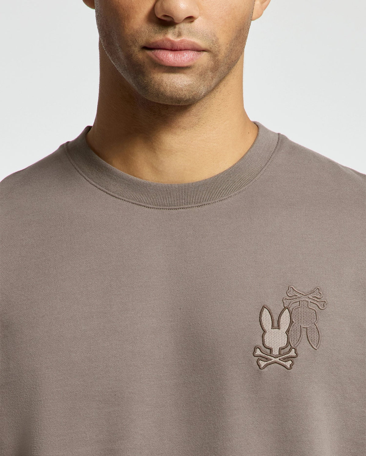 A person wearing the Psycho Bunny MENS PIERCE PEACHED TERRY CREW sweater, stylishly featuring a gray color with an intricate embroidered design of two bunny silhouettes and crossed bones on the chest.