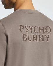 A person wearing a Psycho Bunny Mens Pierce Peached Terry Crew sweater in taupe, featuring "PSYCHO BUNNY" embroidered on the back. Crafted from high-quality cotton, this piece seamlessly combines style and comfort.