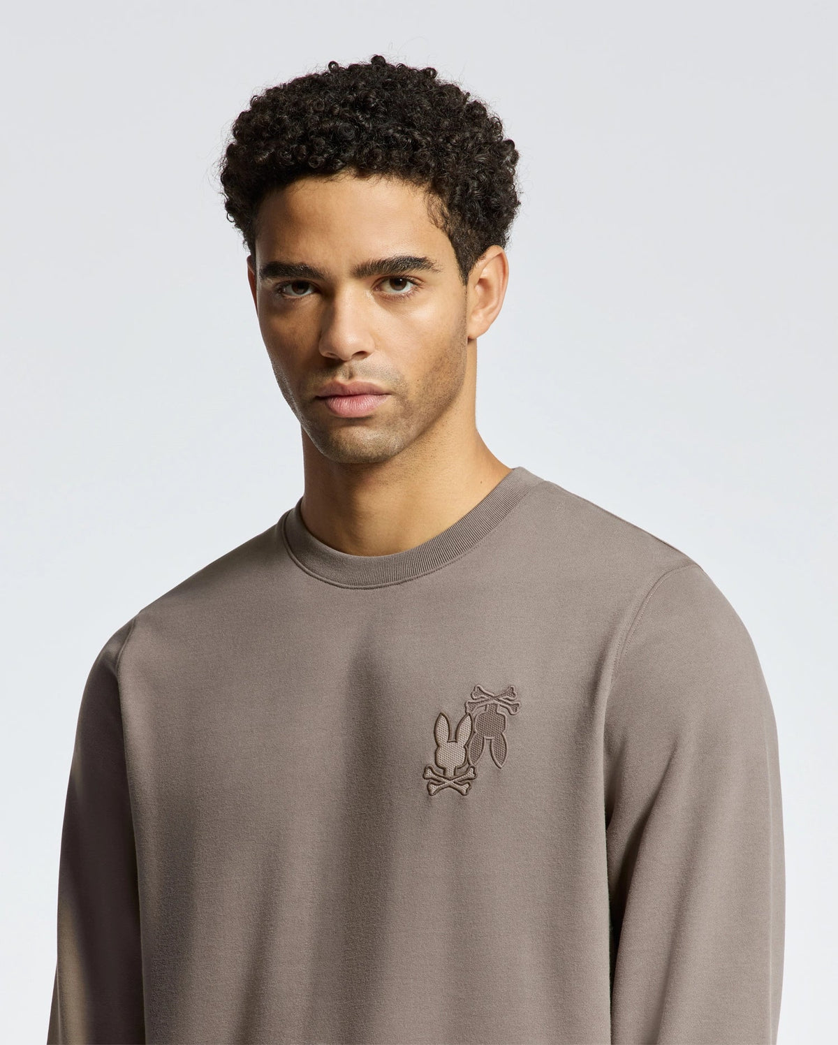 A person stands against a plain background, wearing the Psycho Bunny MENS PIERCE PEACHED TERRY CREW - B6S278E200. The gray sweatshirt features a small chest design and high-quality cotton that offers a simple, casual style.