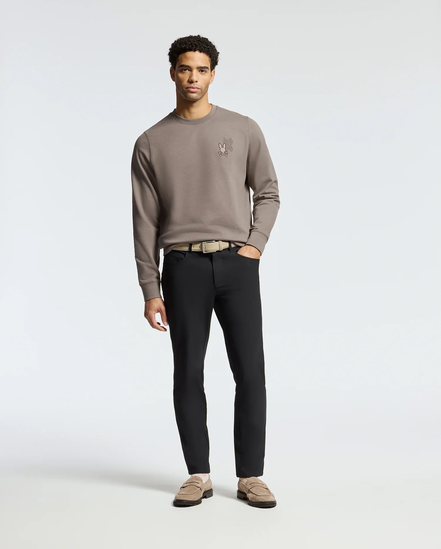 Against a plain background, someone wears the MENS PIERCE PEACHED TERRY CREW - B6S278E200 taupe sweatshirt by Psycho Bunny. Paired with black pants, a beige belt, and brown loafers, their effortlessly stylish look includes relaxed hands by their sides as they gaze ahead.