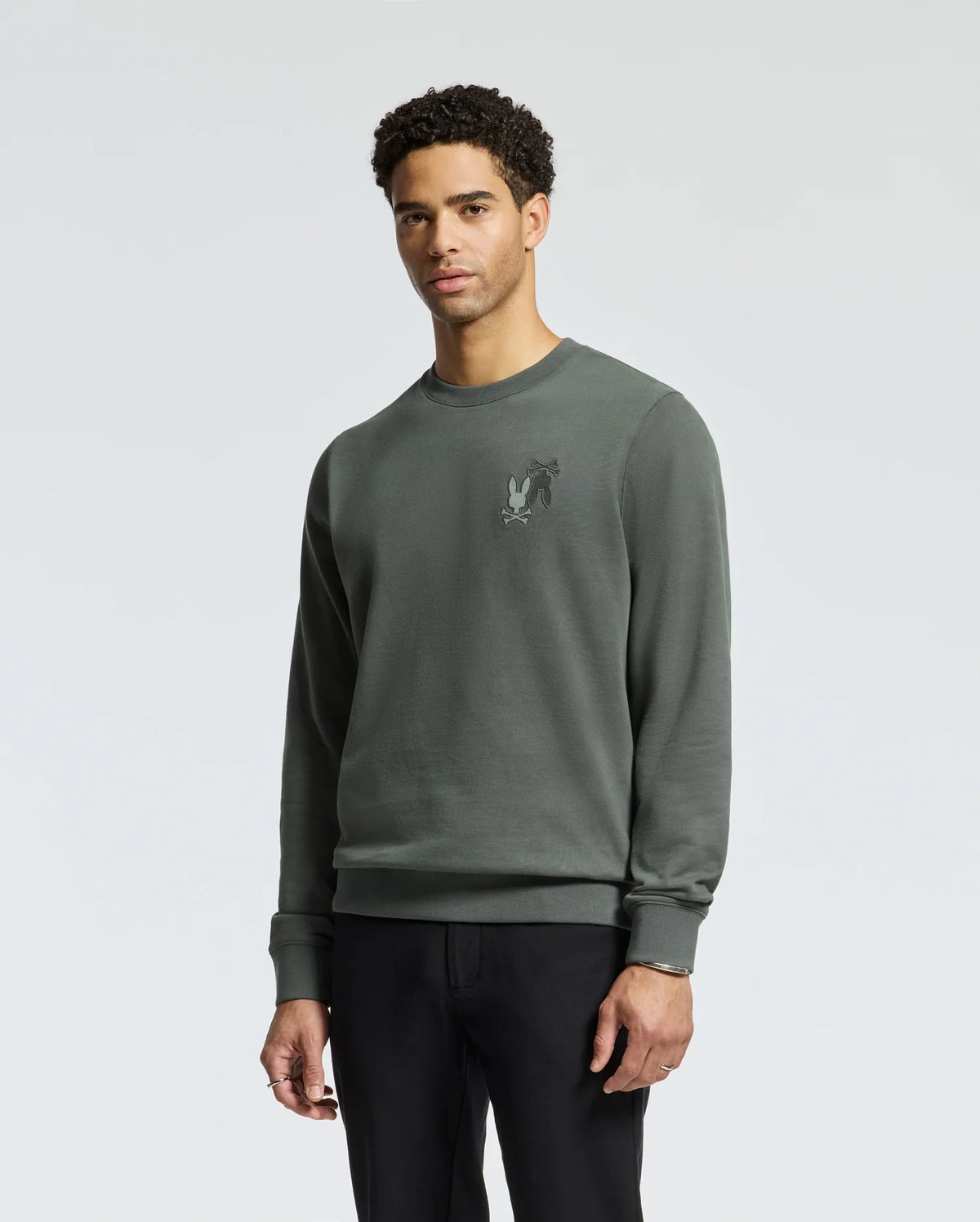 A man with curly hair is wearing the Psycho Bunny MENS WORDMARK PEACHED TERRY CREW - B6S278E200, a dark green knit cotton pullover adorned with embroidered bunnies on the chest. He is standing against a plain light-colored backdrop and has paired it with sleek black pants.