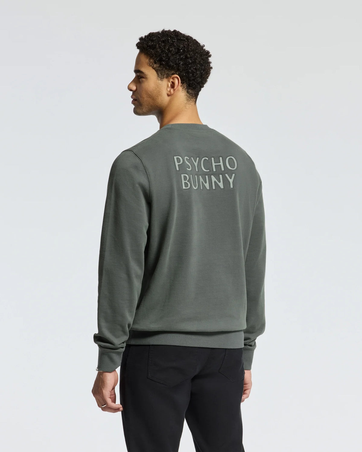 A person with curly hair is turned away, showcasing a dark green Psycho Bunny MENS WORDMARK PEACHED TERRY CREW sweatshirt, featuring "PSYCHO BUNNY" embroidered on the back. They are also wearing black pants against a plain light gray background.