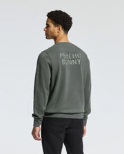 A person with curly hair is turned away, showcasing a dark green Psycho Bunny MENS WORDMARK PEACHED TERRY CREW sweatshirt, featuring "PSYCHO BUNNY" embroidered on the back. They are also wearing black pants against a plain light gray background.