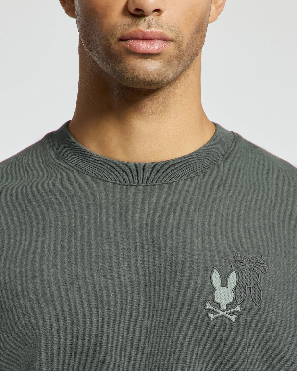 A person is wearing the Psycho Bunny MENS WORDMARK PEACHED TERRY CREW - B6S278E200, a dark green knit cotton crew neck shirt with an embroidered bunny and crossbones logo on the left chest. The image captures them from the shoulders up against a light background.