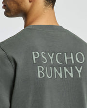A person wearing the Psycho Bunny MENS WORDMARK PEACHED TERRY CREW in dark green, featuring "Psycho Bunny" embroidered in large, textured letters on the back. The knit cotton shirt provides a cozy feel as they turn slightly to the side against a light background.