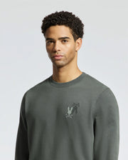 A person wearing the MENS WORDMARK PEACHED TERRY CREW by Psycho Bunny, which is a dark green pullover made of soft knit cotton with a small cartoon print on the left chest, stands against a neutral background. The person has short curly hair and a neutral expression.