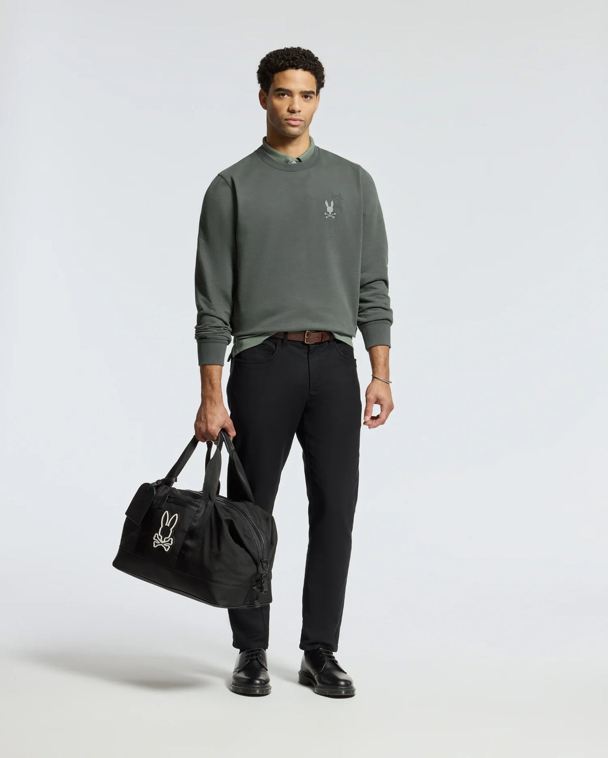 A man in a MENS WORDMARK PEACHED TERRY CREW by Psycho Bunny and black pants holds a sleek black duffel bag featuring an embroidered bunny logo. He stands on a plain white background, confidently facing the camera. With black shoes and short curly hair, he effortlessly embodies style.