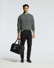 A man in a MENS WORDMARK PEACHED TERRY CREW by Psycho Bunny and black pants holds a sleek black duffel bag featuring an embroidered bunny logo. He stands on a plain white background, confidently facing the camera. With black shoes and short curly hair, he effortlessly embodies style.