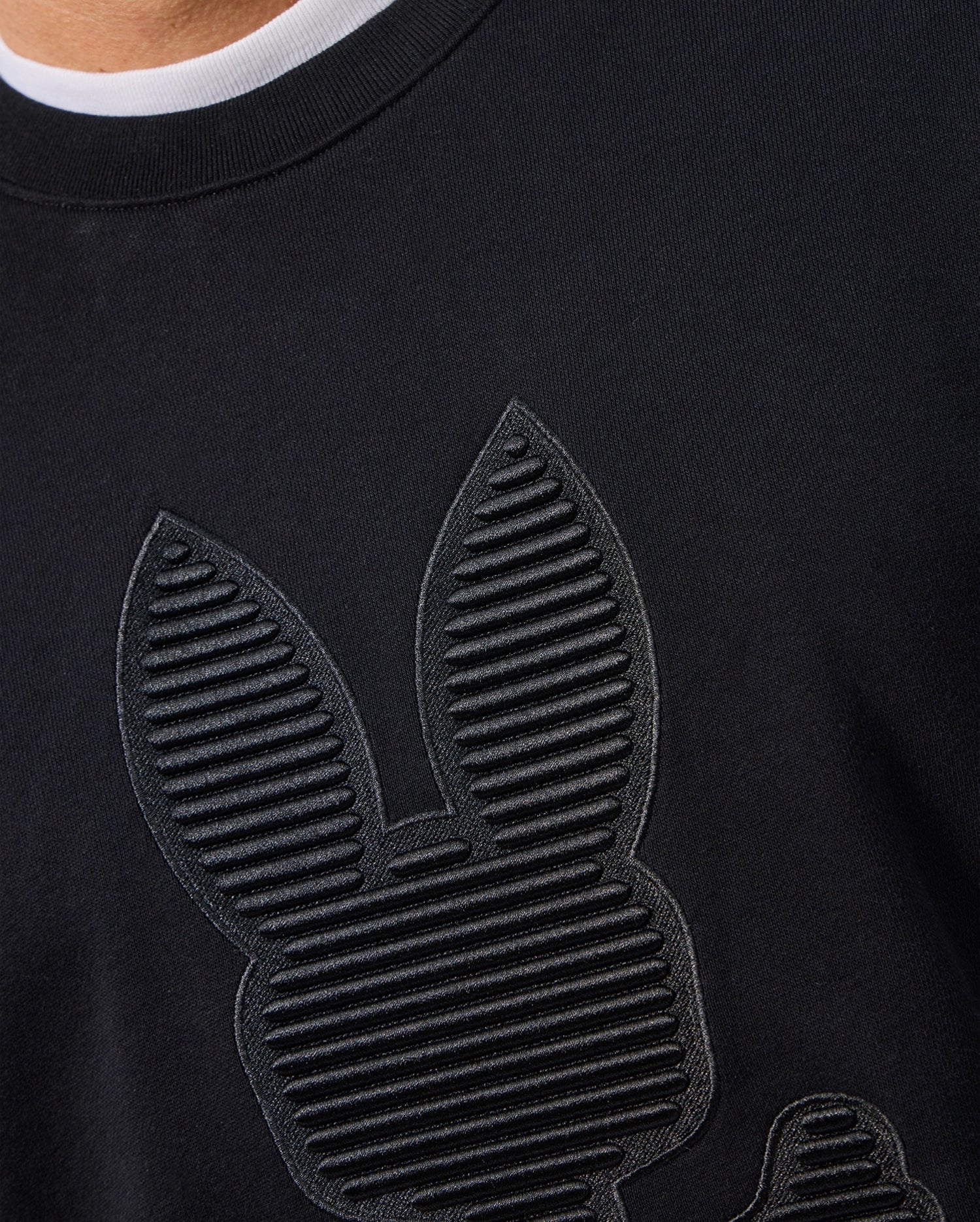 Close-up of a person wearing the MENS KINGSTON SWEATSHIRT by Psycho Bunny. This black cotton jersey shirt features tonal embossing with a textured design showcasing a stylized rabbit head and ears, crafted from horizontal lines to create a remarkable striped effect.
