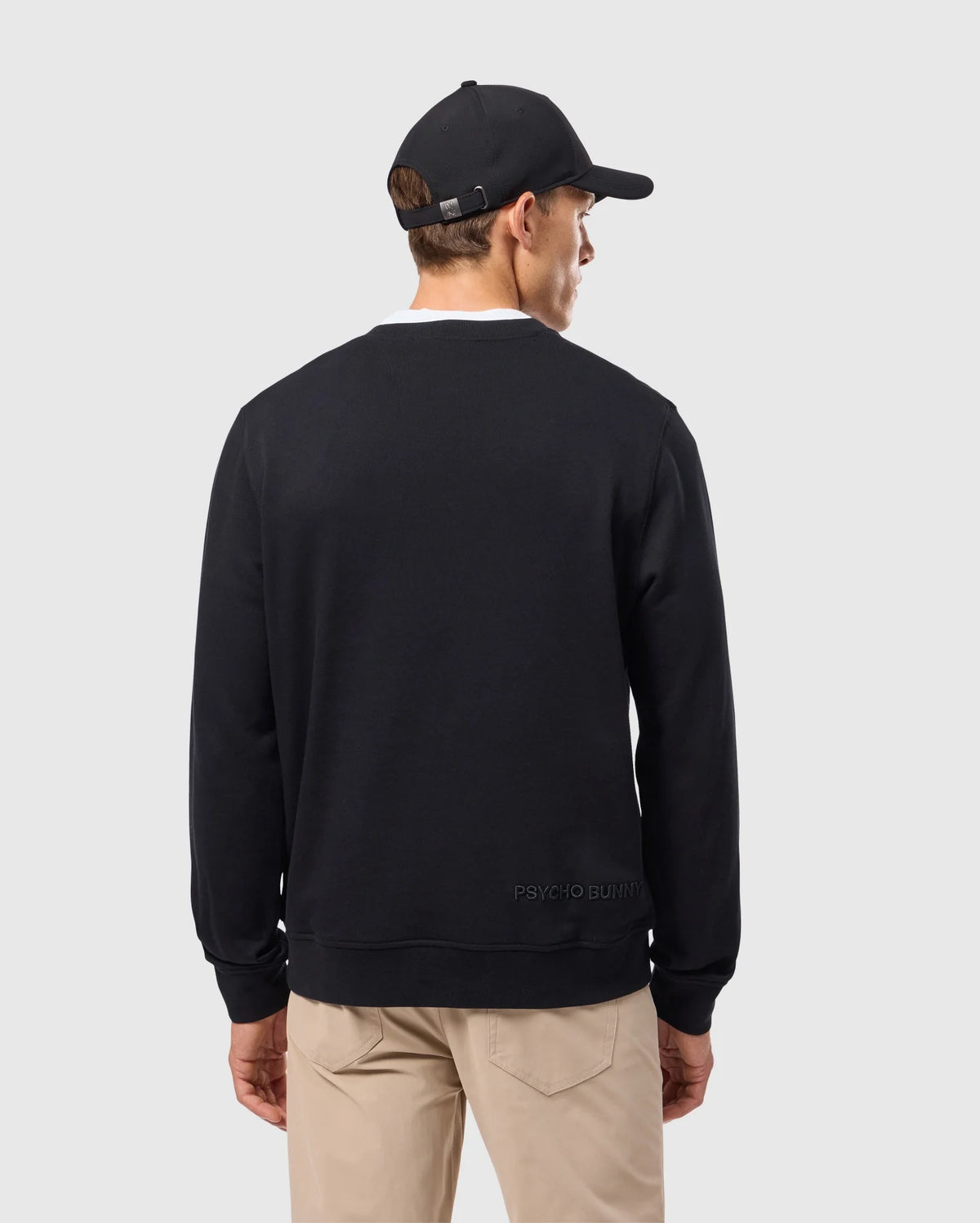 A person wearing a Psycho Bunny MENS KINGSTON SWEATSHIRT - B6S480D200 in black, paired with a black baseball cap and beige pants, stands with their back to the camera against a plain gray background.
