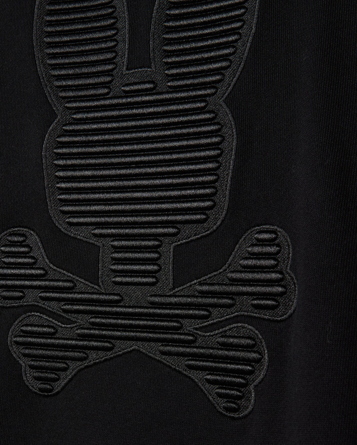 Close-up of the black cotton jersey fabric from the Psycho Bunny MENS KINGSTON SWEATSHIRT (B6S480D200), showcasing a textured design resembling a stylized bunny skull and crossbones. The pattern features tonal embossing with horizontal lines that add detail, creating a 3D-embroidery effect on the sweatshirt.