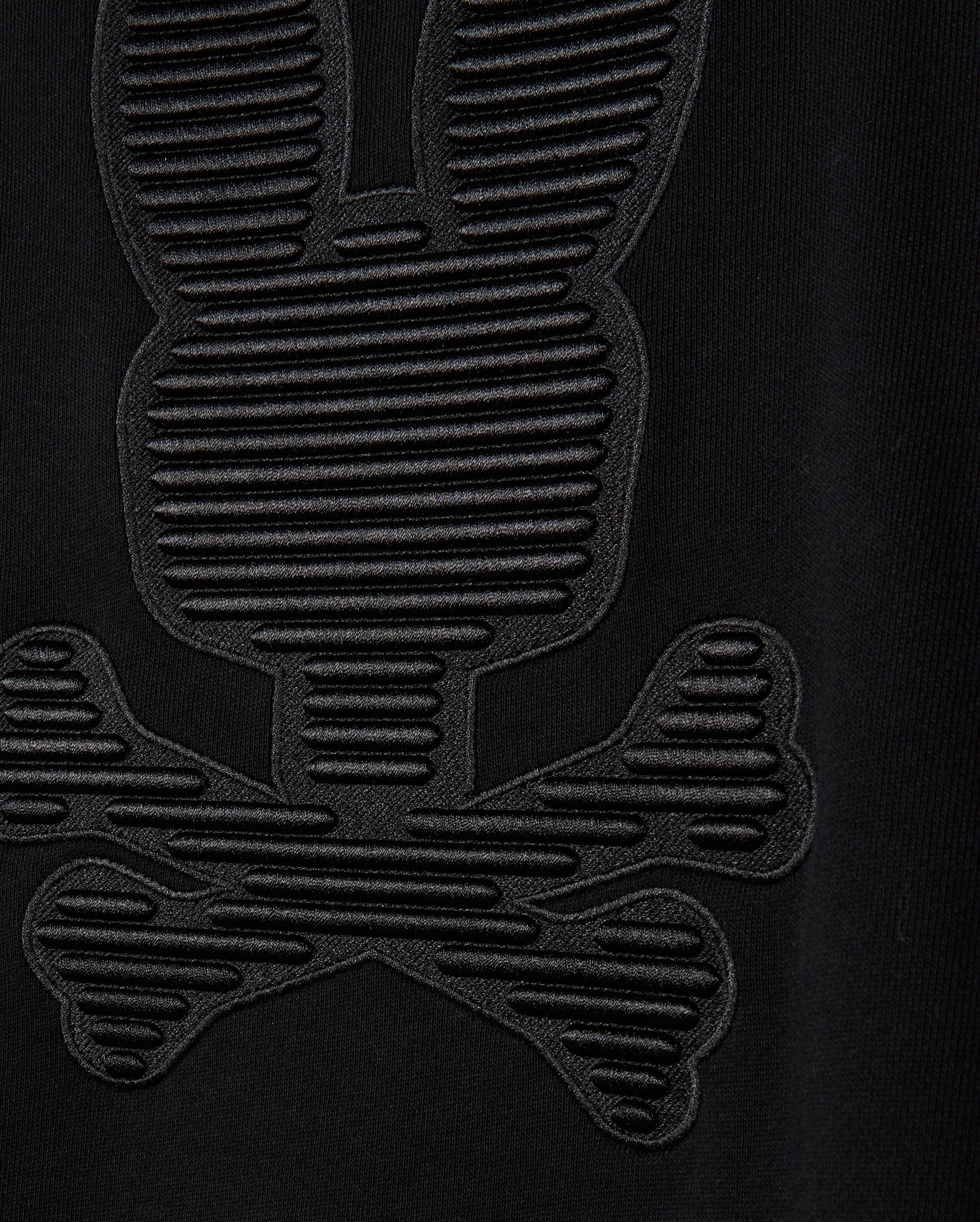 Close-up of the black cotton jersey fabric from the Psycho Bunny MENS KINGSTON SWEATSHIRT (B6S480D200), showcasing a textured design resembling a stylized bunny skull and crossbones. The pattern features tonal embossing with horizontal lines that add detail, creating a 3D-embroidery effect on the sweatshirt.