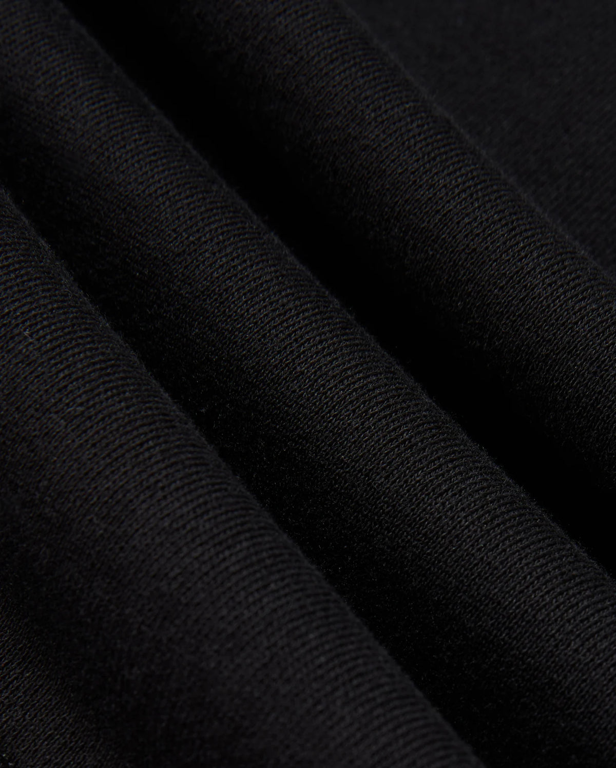 Close-up of the folded black cotton jersey fabric from the Psycho Bunny MENS KINGSTON SWEATSHIRT - B6S480D200, showcasing its fine, textured weave. The folds produce subtle shadows and highlights that enhance the texture and drape of the material, reminiscent of tonal embossing.