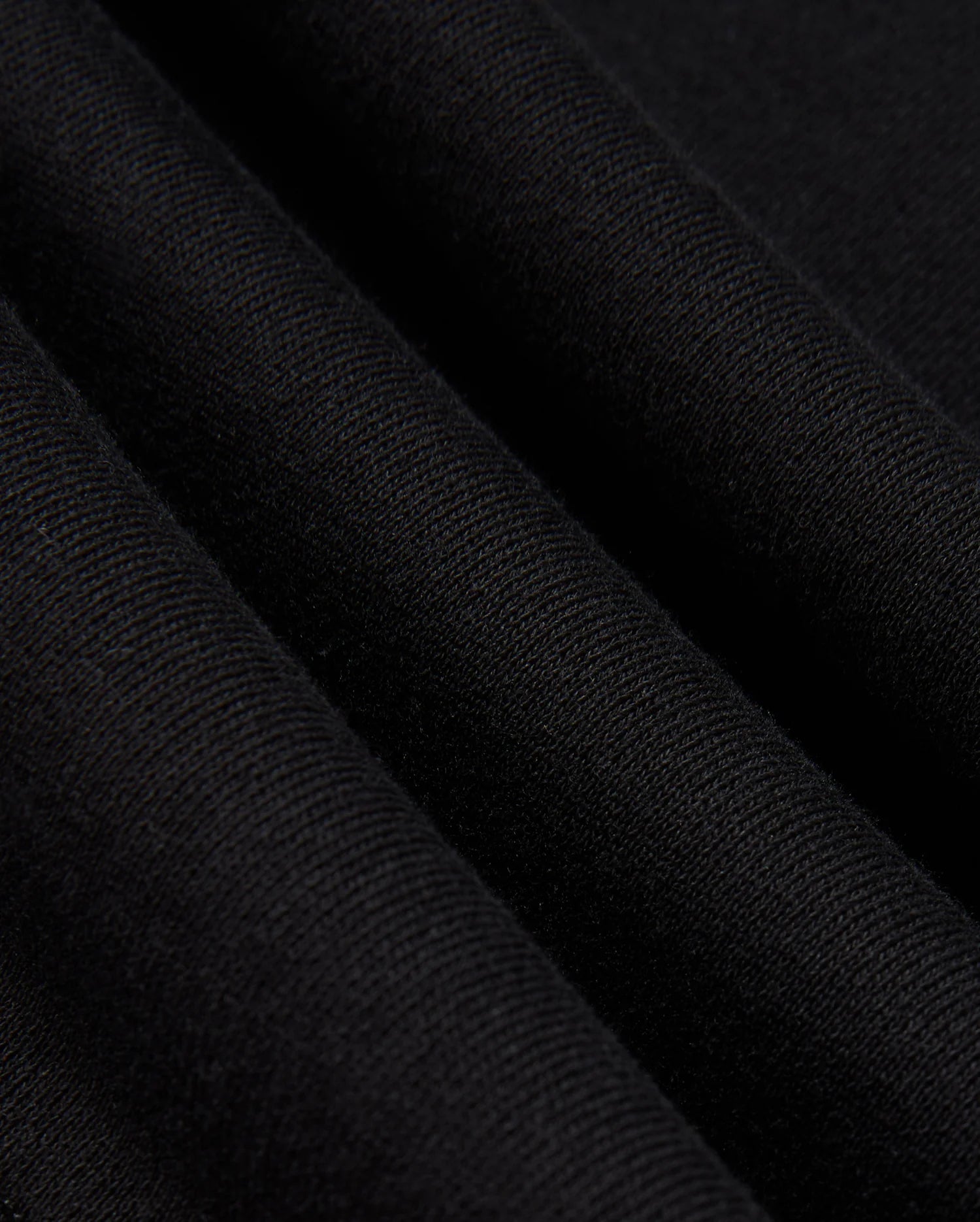 Close-up of the folded black cotton jersey fabric from the Psycho Bunny MENS KINGSTON SWEATSHIRT - B6S480D200, showcasing its fine, textured weave. The folds produce subtle shadows and highlights that enhance the texture and drape of the material, reminiscent of tonal embossing.