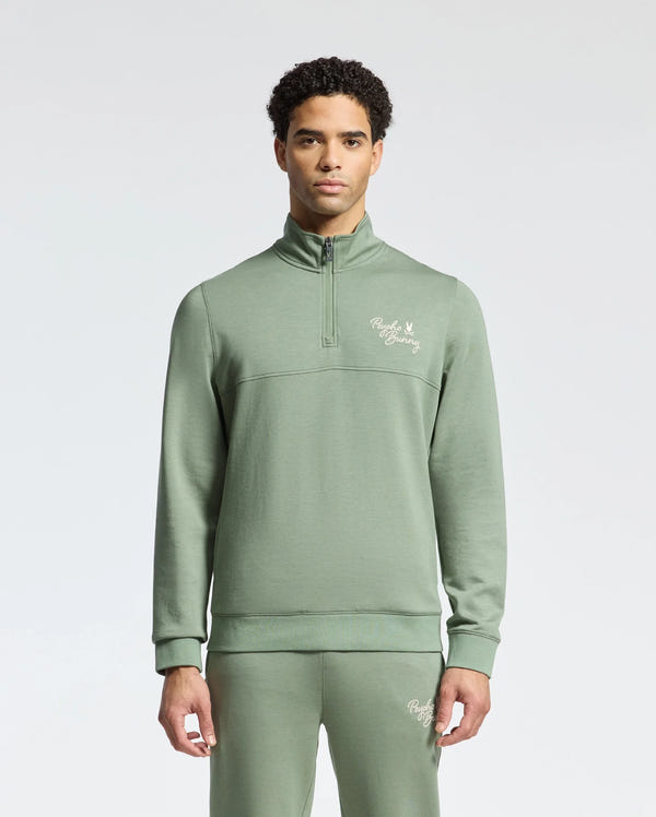 A person is dressed in the Psycho Bunny MENS DESERT 1/4 MOCK ZIP tracksuit, light green with an embroidered wordmark logo on the chest. The 100% cotton set includes a long-sleeved top and matching pants, set against a plain light gray background.