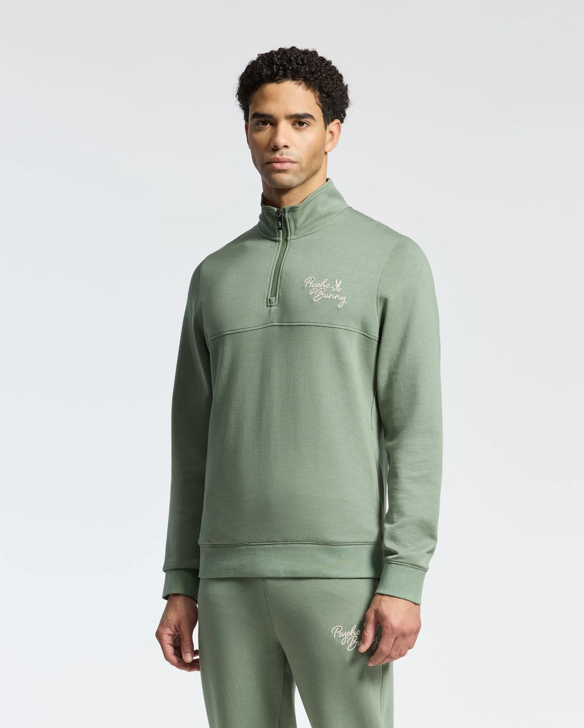 A man in the Psycho Bunny MENS DESERT 1/4 MOCK ZIP - B6S488F200, a light green 100% cotton quarter-zip pullover with matching pants, both with an embroidered wordmark logo on the chest and thigh, stands against a plain white background.